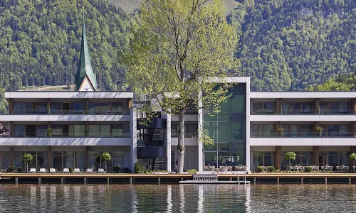 Blick auf ein Seehotel mit alpiner Bergkulisse und Kirchturm – steht für die touristischen Erlebnisse und Gesundheitsangebote der Pletzer Resorts.