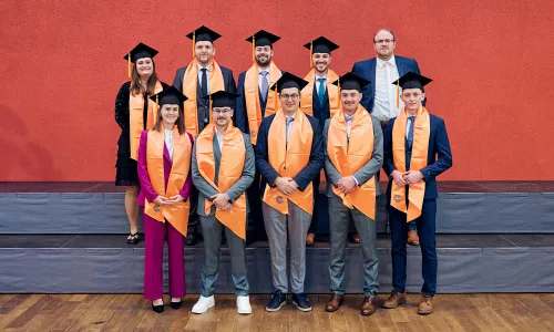 Absolvent:innen in Talaren und orangen Schärpen vor roter Wand – feierlicher Abschluss des dualen Studiums „Smart Building Technologies“ mit Praxisphasen bei iDM Energiesysteme.