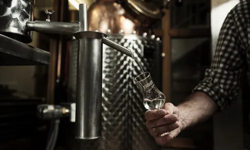 Hand gießt klaren Schnaps aus einer Brennblase in ein Glas – symbolisiert die traditionsreiche Brennkunst der Erber Brennerei in Tirol.